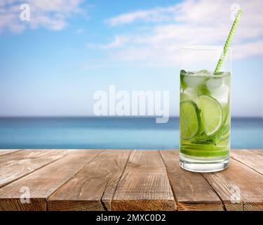 Tasty lemonade with ice cubes and lime on wooden table near sea, space for text Stock Photo
