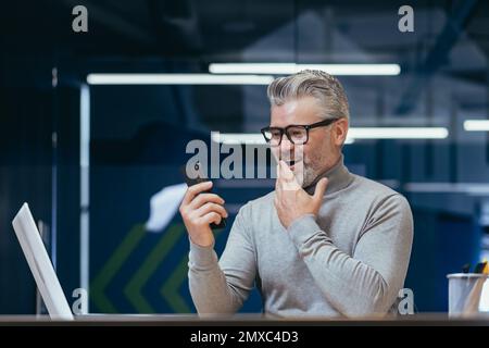 Mature businessman with beard using app on phone inside office, man received online message notification about good news, boss texting online at workplace. Stock Photo