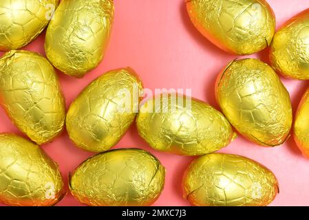 Pile or group of gold, or yellow foil wrapped chocolate easter eggs, against a pink, peach orange background. Stock Photo