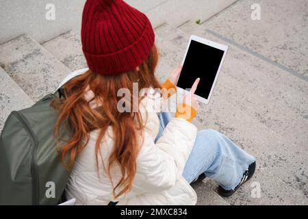 Rear view of redhead girl touches digital tablet screen, touchpad, texts message, uses internet application on gadget, sits on stairs outdoors. Stock Photo