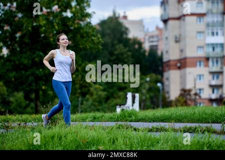 Female runner in white tank top and leggings is jogging in residential area on summer day. Stock Photo