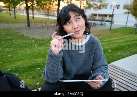 Portrait of asian woman, student in park, sits on bench with digital tablet and a pen, thinking, looking aside thoughtful, making notes, does her Stock Photo