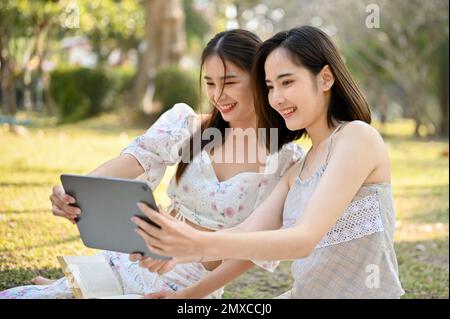 Two beautiful young Asian women are using tablet, taking selfies with tablet or watching videos on a digital tablet together while enjoying a picnic i Stock Photo
