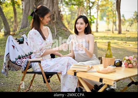 Two beautiful Asian women in lovely dresses enjoying afternoon picnic in the park, sipping white wine, sitting on picnic chairs. Stock Photo