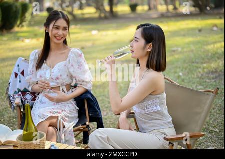 Charming Asian woman in a lovely dress sits on picnic chairs, having a nice conversation, sipping wine, and celebrating a special moment with her frie Stock Photo