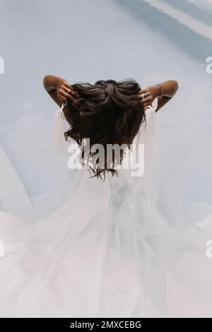 A back view of a bride wearing an elegant white wedding dress while fixing her hair Stock Photo