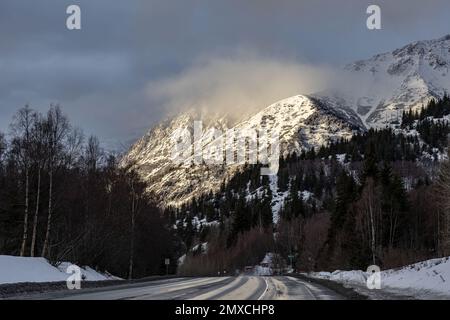 A landscape of a road under snow covered mountains by sunset in Anchorage, Alaska in the winter Stock Photo
