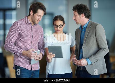 Talking tech in the office. three colleagues talking together over a digital tablet while standing in an office. Stock Photo
