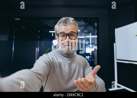Mature businessman having fun talking on video chat using app on smartphone, gray-haired man looking at phone camera and waving hands talking to colleagues while sitting at workplace inside office. Stock Photo
