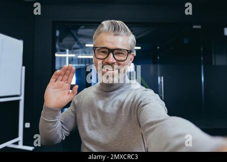 Mature businessman having fun talking on video chat using app on smartphone, gray-haired man looking at phone camera and waving hands talking to colleagues while sitting at workplace inside office. Stock Photo