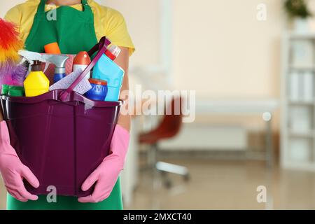 Woman holding bucket with cleaning supplies in office, closeup. Space for text Stock Photo