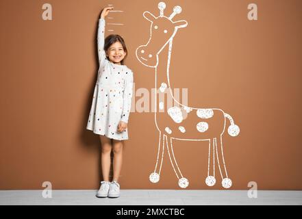 Little girl measuring height and drawing of giraffe near brown wall Stock Photo