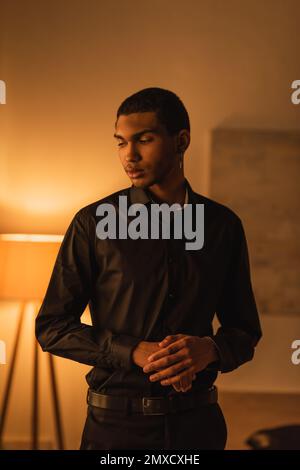 brunette african american man in black stylish clothes standing in dark room with luminous lamp Stock Photo