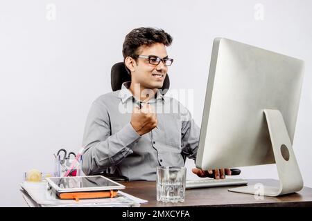 A smiling young Indian businessman in formal clothes clenching his fist while looking at the computer Stock Photo