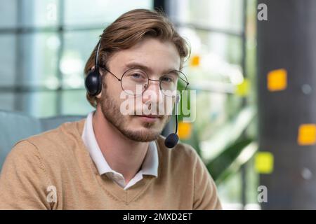 Close-up photo. Portrait of a young handsome man in a headset, glasses and a beard. He works in the office at a desk. He looks seriously into the camera. Stock Photo