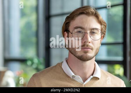 Close-up photo. Portrait of young handsome man freelancer, programmer, designer blond in glasses and beige sweater with shirt. He looks seriously into the camera. Stock Photo