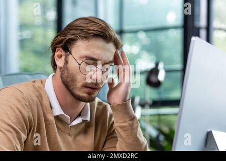 Close-up photo. Young man freelancer, office worker sits in the office at the table, holds and massages his head with his hand, closed his eyes, feels a severe headache. Working overtime, deadline. Stock Photo