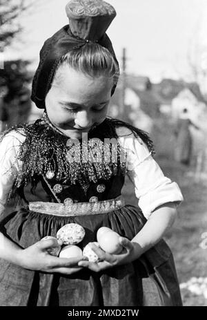 Mädchen in Tracht mit gefundenen Ostereiern, Schwalm-Eder-Kreis in Hessen, 1938. Girl in traditional costume with found Easter eggs, Schwalm-Eder region in Hesse, 1938. Stock Photo