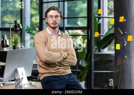 Portrait of a young handsome man freelancer, programmer, He sits on the desk in the office, crosses his arms, looks seriously into the camera. Stock Photo