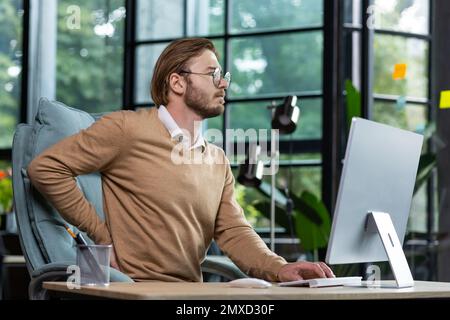 Health problems from sedentary work. A young man businessman is tired at the workplace in the office, feels pain in his back and body, massages his back with his hand, works hard, overtime work. Stock Photo