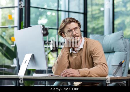 Overtime work. Deadline. A young man businessman sits tired in the office at the table. Feels pain in the body, massages the neck with his hand, rests, took a break. Stock Photo