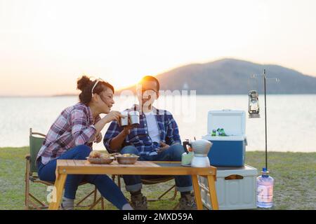 Asian couple having a cup of coffee or water at their camping site with lake in the background during sunset Stock Photo