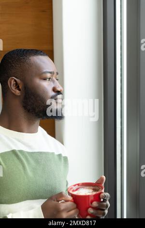 Vertical of thoughtful african american man with cup of coffee looking out of window, copy space Stock Photo