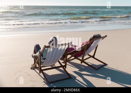 African american senior couple talking while sitting on deckchairs in front of seascape at beach Stock Photo