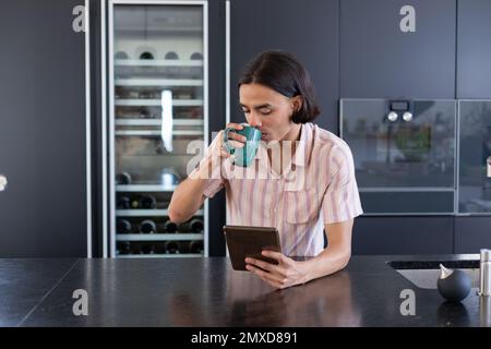 Happy biracial man using tablet and drinking coffee in kitchen Stock Photo