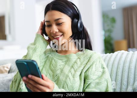 Image of happy biracial woman in headphones using smartphone sitting on sofa at home, copy space. Leisure, technology and domestic life concept. Stock Photo