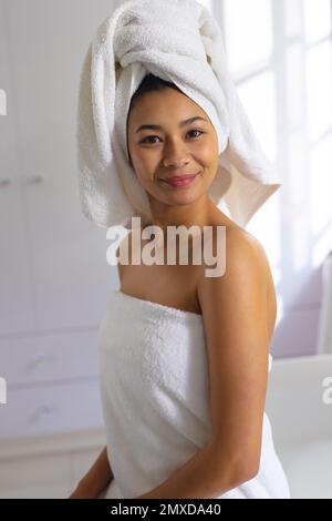 Vertical portrait of happy biracial woman wearing towel, smiling in bathroom, with copy space. Relaxation, self care, beauty, leisure and lifestyle co Stock Photo