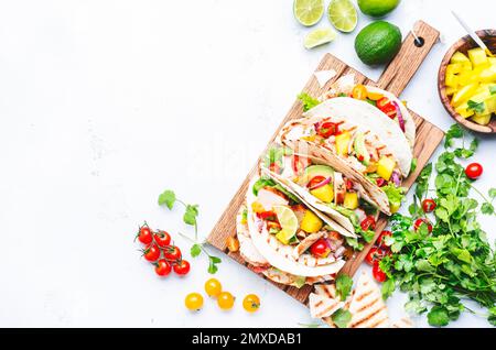 Tacos with grilled chicken, tomato salsa sauce, mango, cilantro and onion in tortilla on cutting board. White table background, top view Stock Photo