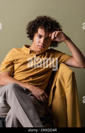 trendy african american man in yellow polo shirt sitting with crossed legs and hand near head on grey background,stock image Stock Photo