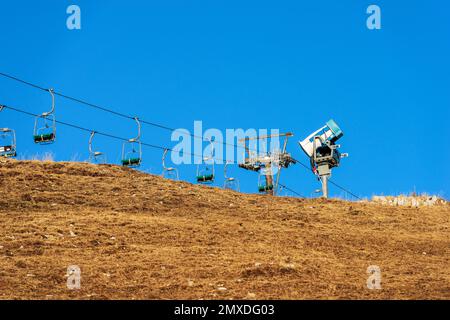 Snow cannon or snowmaking system and an empty chairlift in winter on a brown meadow, ski slope, without snow due to the too hot. Veneto, Italy. Stock Photo