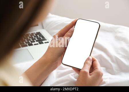 Young woman holding mobile phone with empty screen in hands on bed at home, closeup Stock Photo