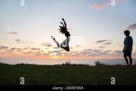 silhouette of teenager girl joyfully bounces, has fun at sunset by sea. concept of freedom, happiness. girl trains with pleasure, guy or coach looks a Stock Photo