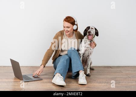 Smiling redhead woman in headphones using laptop and hugging dalmatian dog at home,stock image Stock Photo