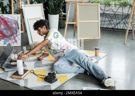 Side view of african american artist holding smartphone and using record player near books and vintage camera in studio,stock image Stock Photo