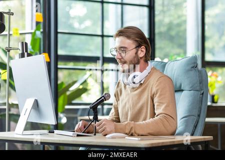 Young handsome male blogger sitting at the desk in the office at the computer in front of the microphone. Records a podcast, talks on a video call, communicates in an online chat. Stock Photo