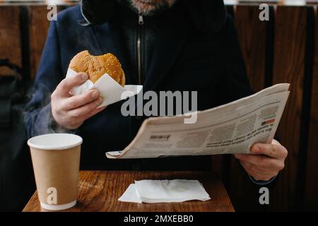 A middle-aged bearded man eats a tasty burger and reads today's newspaper on a city street in winter. Street food concept. Stock Photo