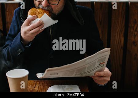 A middle-aged bearded man eats a tasty burger and reads today's newspaper on a city street in winter. Street food concept. Stock Photo