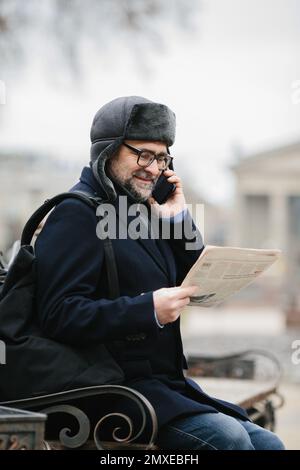 An elderly man enjoys a cold winter morning in the city. A man sits on a bench in the square, reads a newspaper and cheerfully chats with friends on a Stock Photo