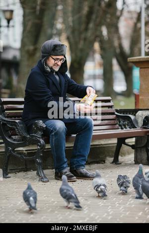 An elderly man enjoys a cold winter morning in the city. A gray-haired man feeds pigeons while sitting on a bench in the square. Stock Photo