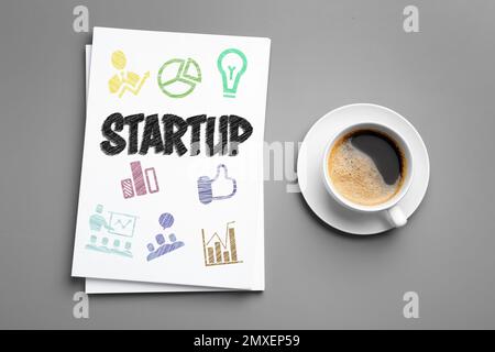 Paper sheets with word STARTUP and drawn colorful icons near cup of coffee on grey background, flat lay Stock Photo