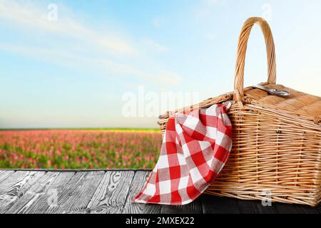 Wicker picnic basket with checkered tablecloth on wooden table in field, space for text Stock Photo