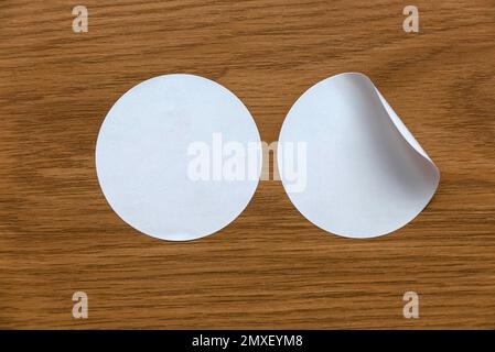 Blank white round stickers straightened and with folded corner on wooden background. Stock Photo