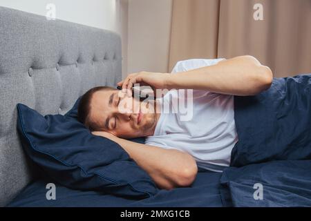 Happy young cheerful man in pajamas wearing a sleep mask. Rest, rest at home, sit, turn around under a blanket, blanket, point a finger at a mobile ph Stock Photo