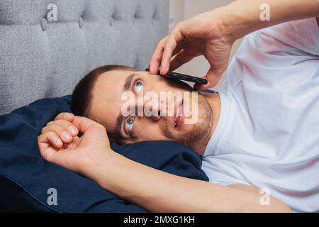 Happy young cheerful man in pajamas wearing a sleep mask. Rest, rest at home, sit, turn around under a blanket, blanket, point a finger at a mobile ph Stock Photo