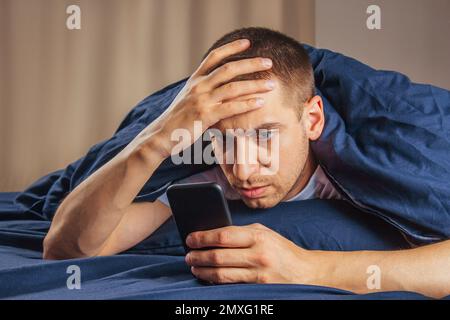 Bad news, problems, offers and advertising. Shocked upset attractive guy reads a message on the phone and presses his hand to his face on a white bed Stock Photo