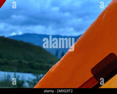Raindrops lie on the tent of an orange tourist tent against the backdrop of a mountain and a river. Stock Photo
